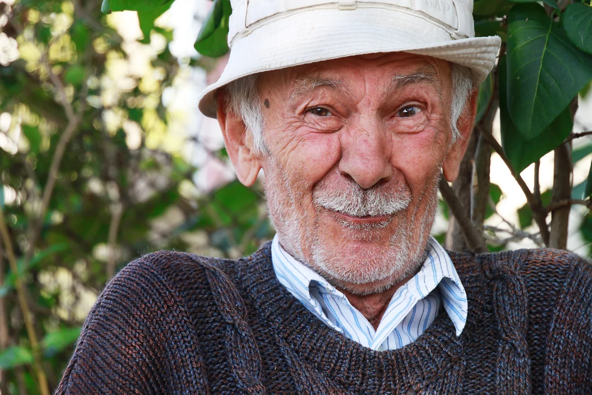 Award-winning portrait photograph of an elderly man in a bucket hat, smiling warmly among green foliage.